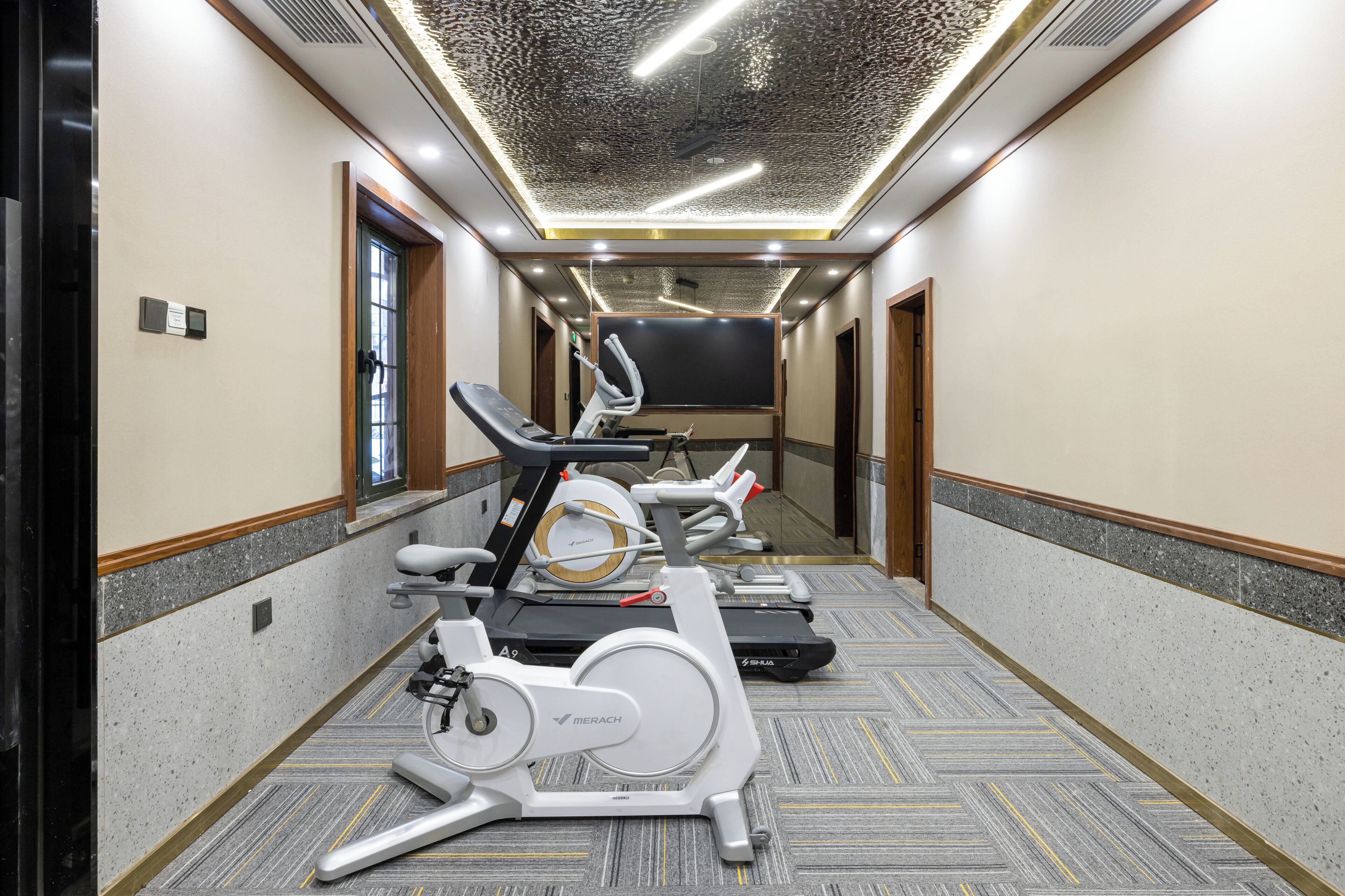 A hotel corridor with exercise equipment, including treadmills and stationary bikes, and a large flat-screen TV mounted on the wall.
