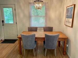 A dining area with a wooden table and four grey upholstered chairs, a chandelier, a window with blinds, and a framed picture on the wall.