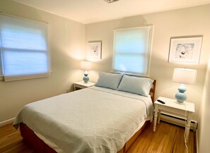 A bedroom with a bed, two bedside tables with lamps, a window with blinds, and a framed picture on the wall.