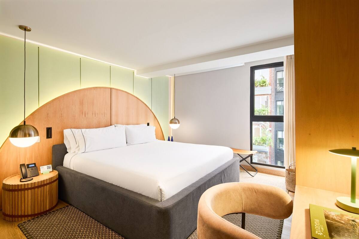 Un hotel moderno con una cama grande, lámparas de noche, una mesita redonda, una silla y vistas al exterior por una ventana.