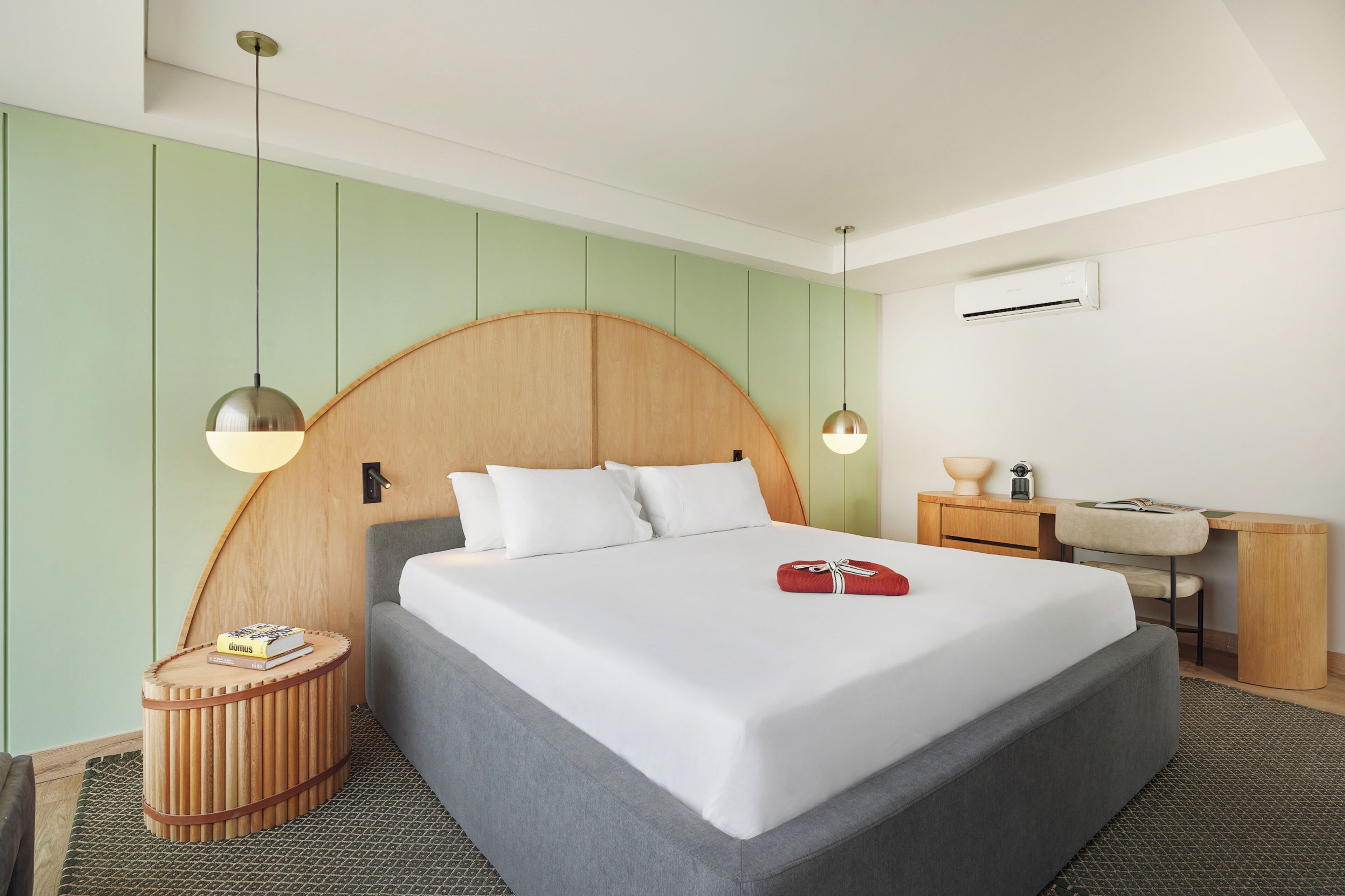 A modern hotel room with a large bed, a wooden headboard, a round wooden side table, a desk with a chair, and a small round rug.