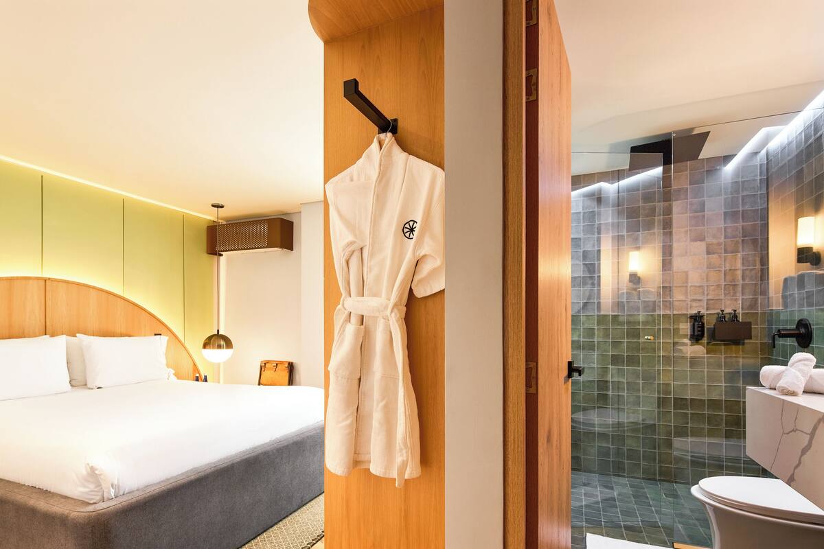 Una habitación de hotel con cama, una bata colgada en un perchero, cabecera de madera, mesita de noche y baño con ducha e inodoro.