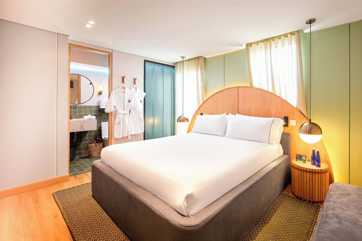 Habitación de hotel con una cama grande, una mesita de noche de madera con lámpara, un baño con espejo y toallero, y una pared verde.