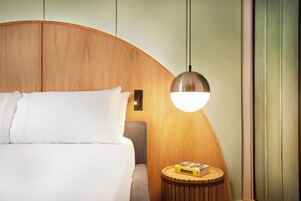 Un dormitorio con un cabecero de madera curvada, una cama con ropa de cama blanca, una mesita de noche redonda de madera con un libro, y una lámpara colgante moderna.