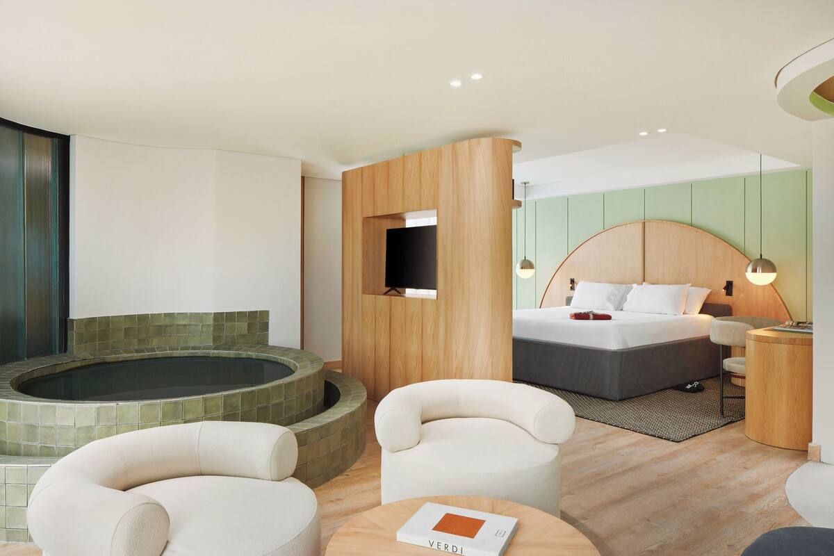 Un hotel moderno con una cama grande, una zona de estar con sillas, una mesita con un libro, una bañera y un cabecero de madera.