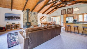 A spacious living room with a large leather sofa, a stone fireplace, wooden beams, a kitchen with a bar area, and a view of the outdoors. - Ski Chalet|Ping-Pong|Fire Pit|5 min -> Slopes 🎿❄️🏂🌲🗻 (Bartlett)