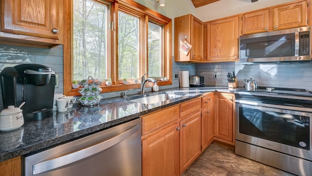 A modern kitchen with wooden cabinets, stainless steel appliances, a large window, and a countertop with various items. - Ski Chalet|Ping-Pong|Fire Pit|5 min -> Slopes 🎿❄️🏂🌲🗻 (Bartlett)