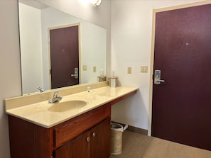 A bathroom with a double sink, a large mirror, a wooden vanity, a trash can, and a door.