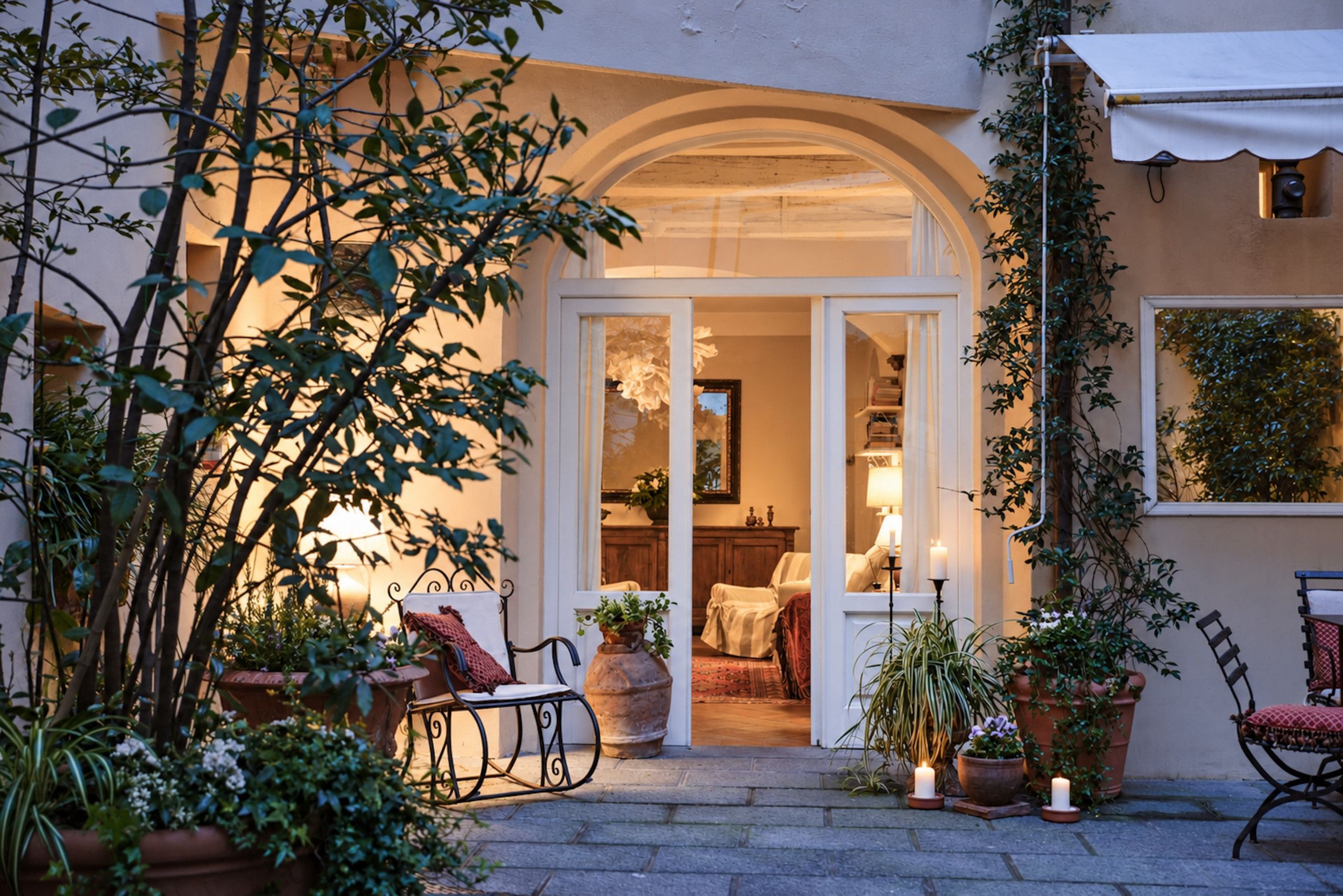 A cozy outdoor seating area with a wrought iron chair, potted plants, and a doorway leading to an interior space with a lamp and a chair.