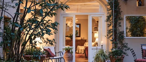 A cozy outdoor seating area with a wrought iron chair, potted plants, and a doorway leading to an interior space with a lamp and a chair.