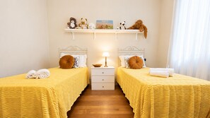 A hotel room with two beds, a nightstand with a lamp, a small shelf with decorative items, and a window with curtains.