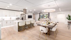 A modern kitchen with white cabinetry, a central island with bar stools, and a dining area with a wooden table and white chairs. - The Rody - Mins to Old Town with Heated Pool, Games, Fast Wifi and More... (Scottsdale)