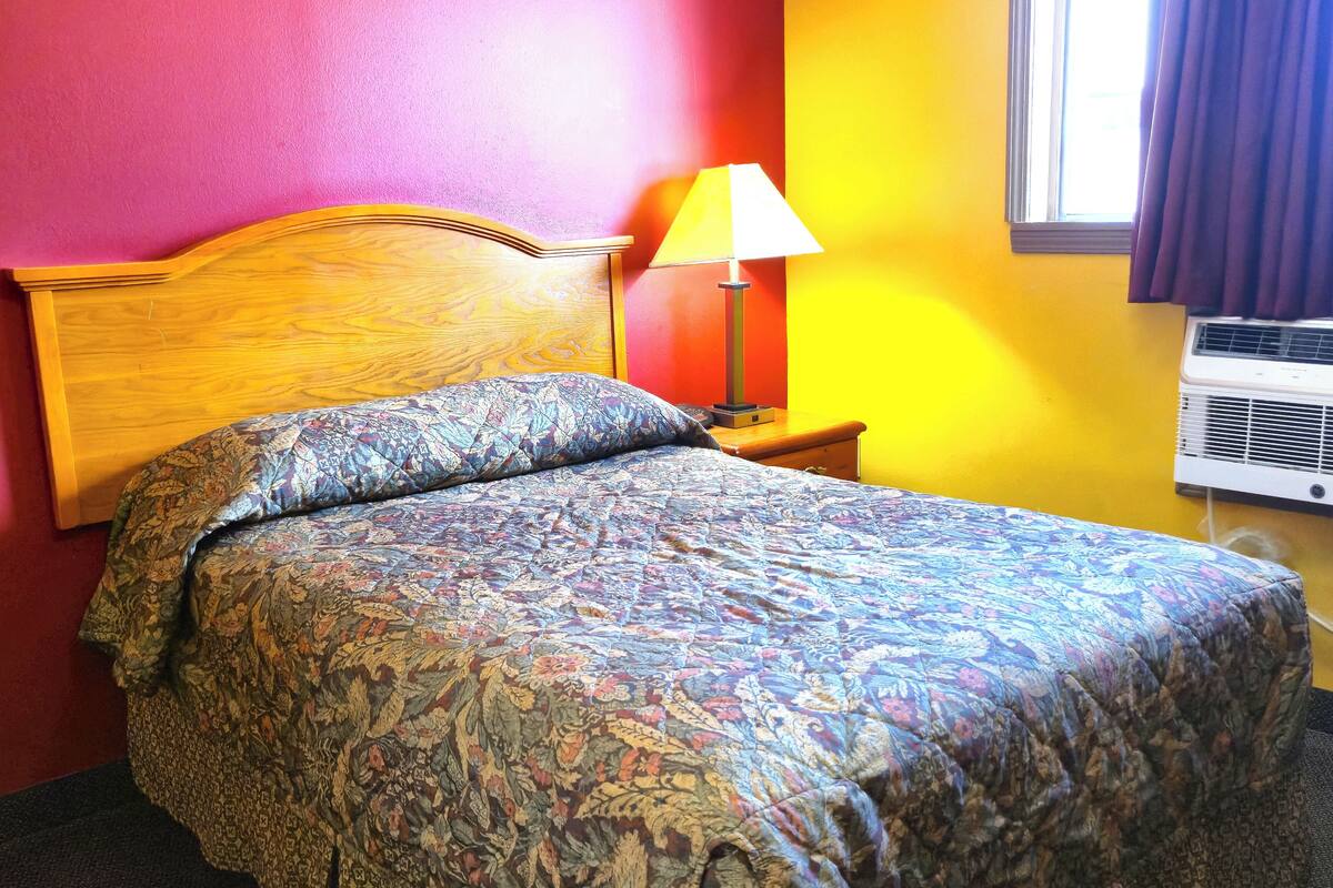 Un lit avec une couvre-lit à motifs floraux, une tête de lit en bois, une table de chevet avec une lampe et une fenêtre avec des rideaux.