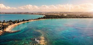 An aerial view of a coastal area with clear turquoise waters, a sandy beach, and a densely forested area.