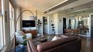 Un salon spacieux avec un grand canapé en cuir marron, un téléviseur à écran plat fixé au mur et un coin repas comprenant une table et des chaises en bois.