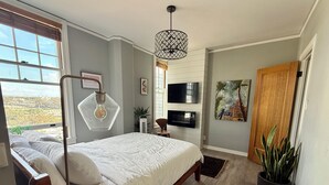 Une chambre avec un grand lit, un téléviseur fixé au mur, une plante en pot et une vue sur l’extérieur.