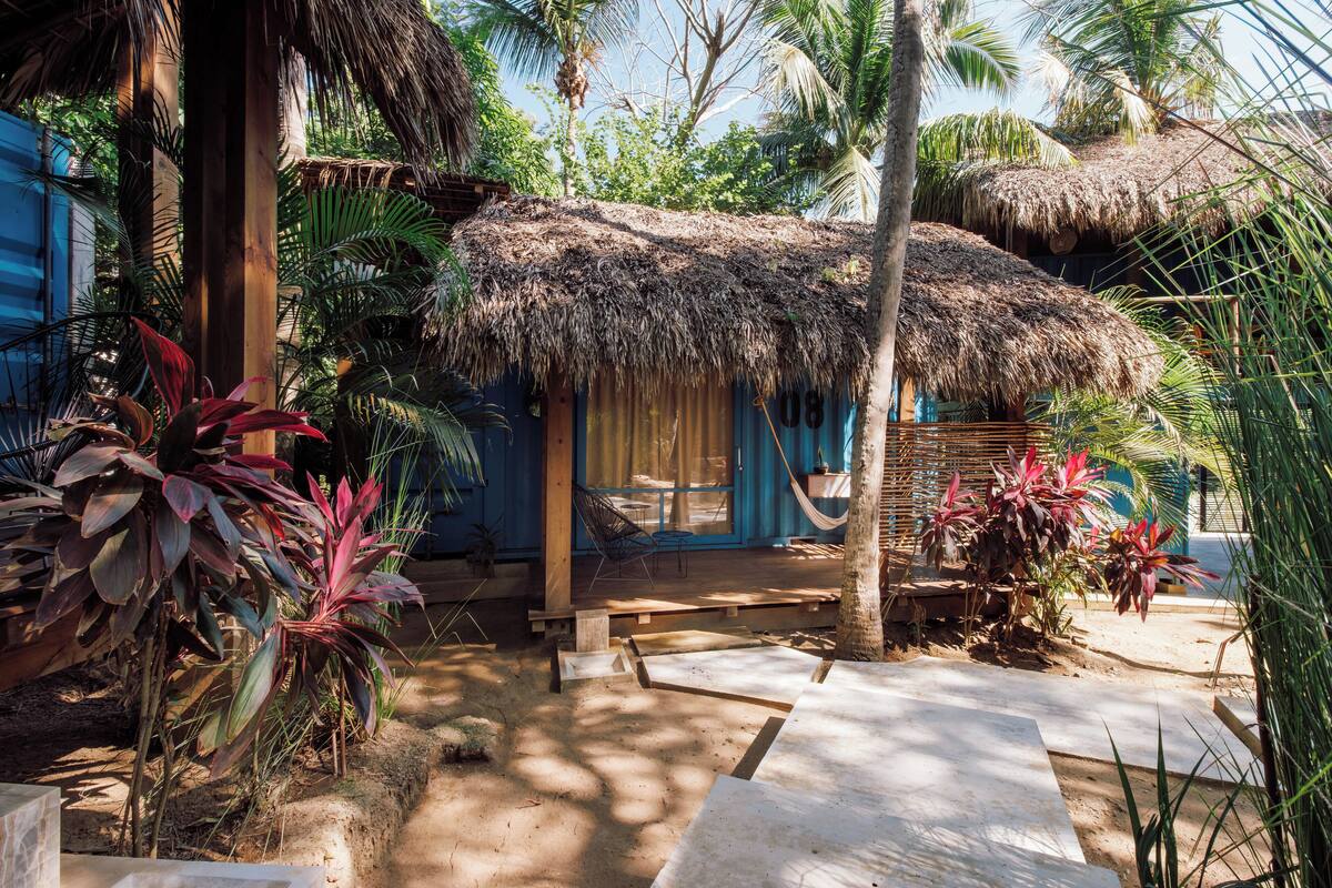 Un espacio al aire libre rústico con una construcción de techo de paja, rodeado de plantas tropicales y un banco de madera.