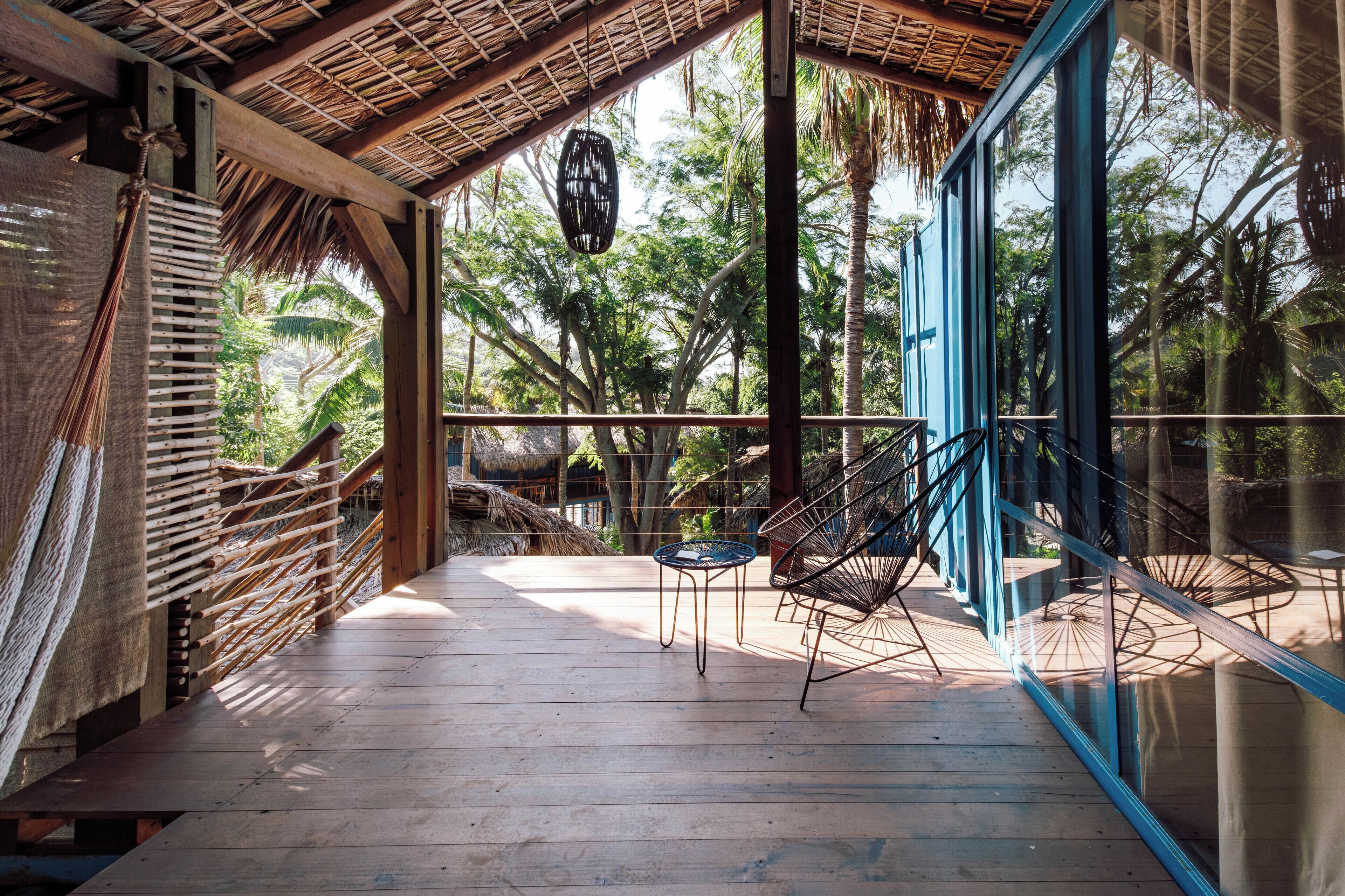 Une terrasse en bois surmontée d’un toit de chaume, une porte vitrée à cadre bleu et un luminaire suspendu.
