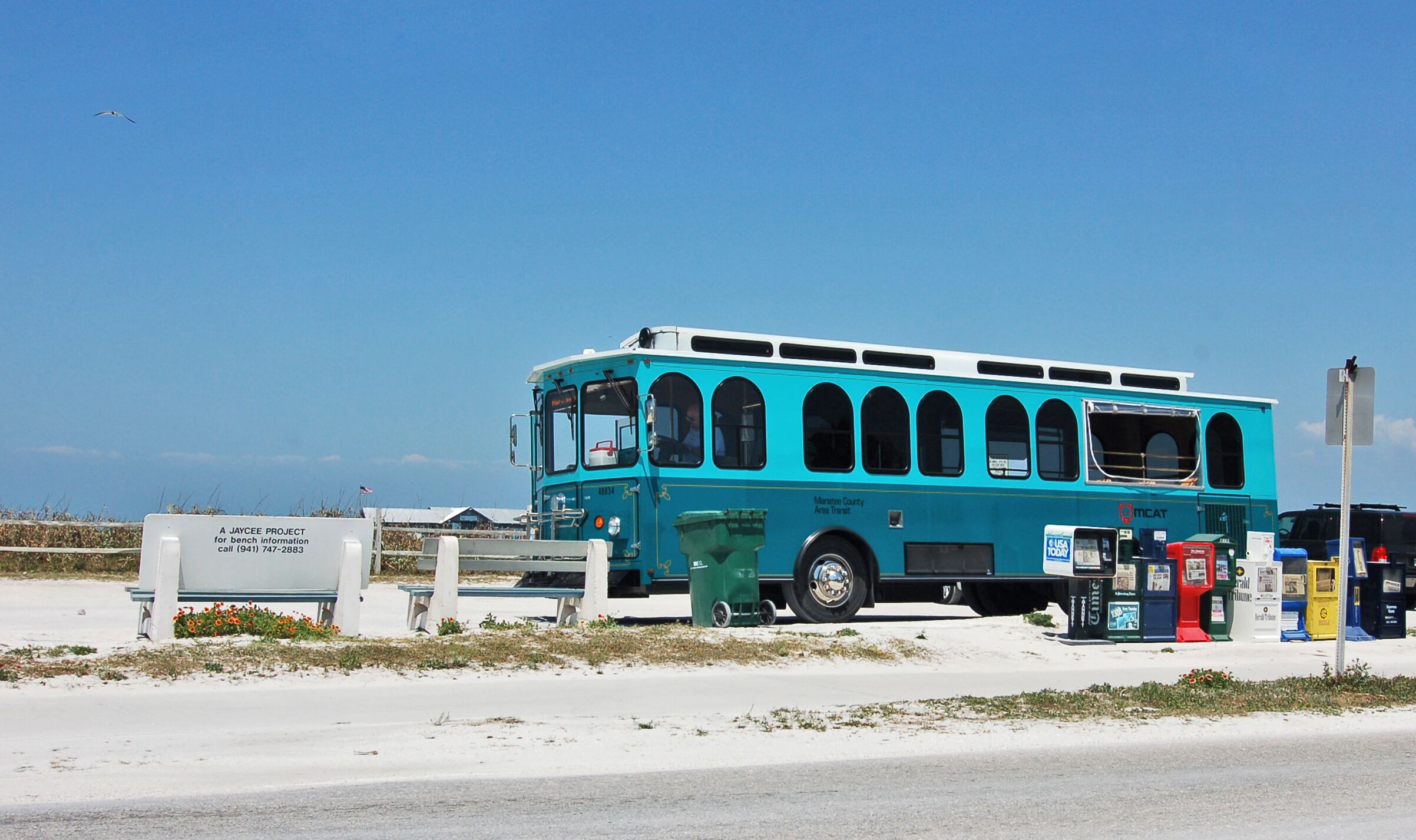 A turquoise bus parked on a sandy area with a bench and trash bins nearby.