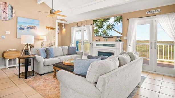 A living room with a sofa, coffee table, and a fireplace. There is a ceiling fan, a painting, and a sign that reads "BEACH HOUSE".