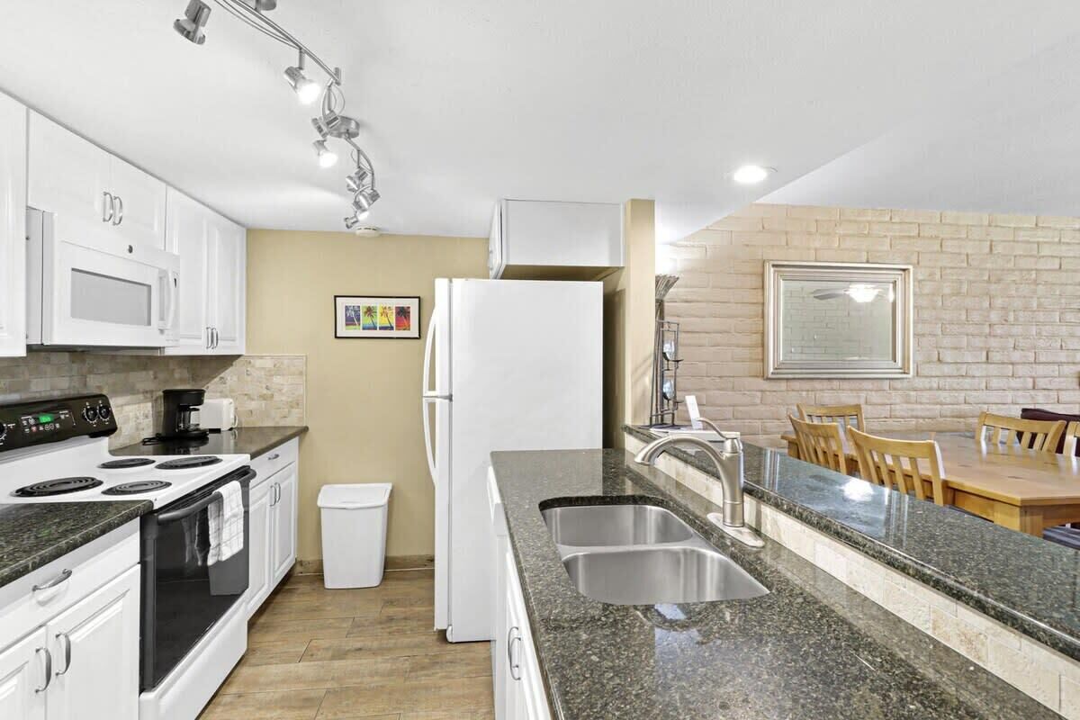A modern kitchen with white cabinets, a granite countertop, a stainless steel sink, and a dining area with a brick wall and wooden table.