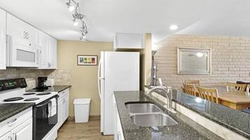 A modern kitchen with white cabinets, a granite countertop, a stainless steel sink, and a dining area with a brick wall and wooden table.