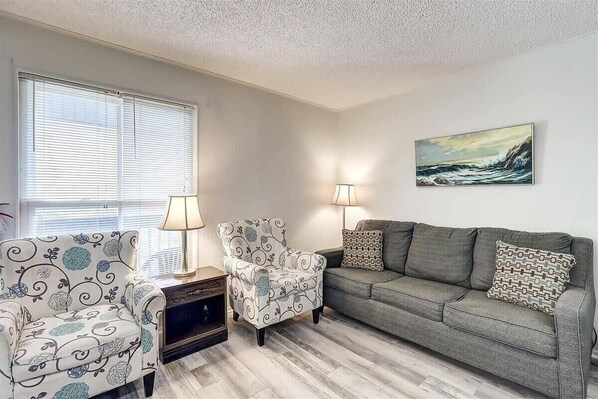 A living room with a grey sofa, patterned armchairs, and a painting on the wall. - Pompano- 2 Min Drive to Beach! | Sleeps 4 (Port Aransas)