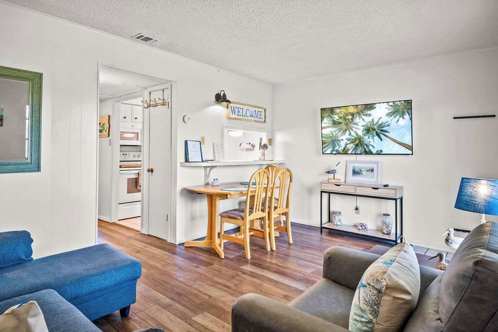 A living room with a blue sofa, a grey sofa, a wooden dining table with chairs, a TV, and a 'WELCOME' sign on the wall.