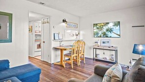 Un salon avec un canapé bleu, un canapé gris, une table à manger en bois avec des chaises, un téléviseur et un panneau « BIENVENUE » accroché au mur.