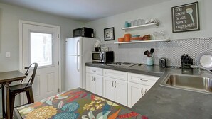 A kitchen with white cabinets, a microwave, a refrigerator, and a sink. There is a colorful tablecloth on the table, and a shelf with dishes and glasses. - Kona Cottage~Dog Friendly~In Town Location (Port Aransas)