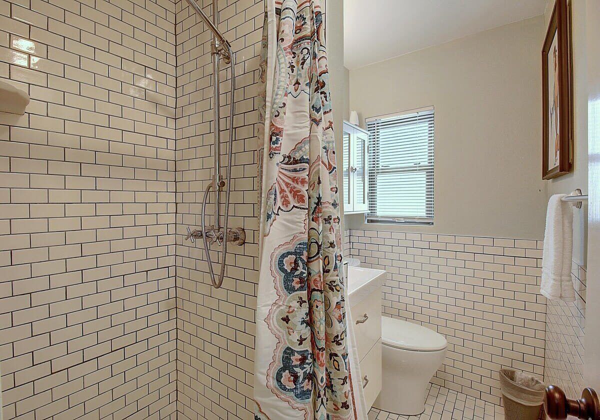 A bathroom with white tiled walls, a shower with a patterned curtain, a toilet, and a window with blinds.