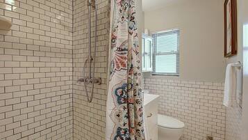 A bathroom with white tiled walls, a shower with a patterned curtain, a toilet, and a window with blinds.