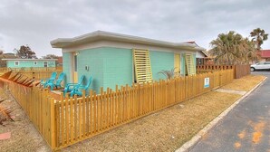 A small, single-story building with a light blue exterior and yellow shutters, surrounded by a wooden fence. - Kona Cottage~Dog Friendly~In Town Location (Port Aransas)