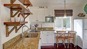 A kitchen with granite countertops, white cabinets, wooden shelves, a sink, and a dining area with two wicker chairs.