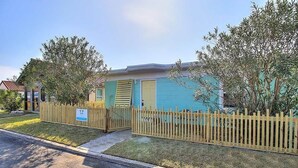 Une maison de plain-pied à la façade bleu clair, avec une clôture en bois et un petit jardin devant, composé de pelouse et d’arbres.