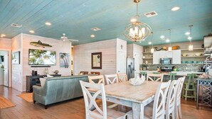 Un vaste espace de vie comprenant une salle à manger avec une table en bois, des chaises blanches, un canapé et une cuisine équipée d’appareils électroménagers modernes.