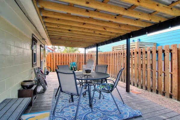 An outdoor patio with a wooden deck, metal table and chairs, and a blue patterned rug.