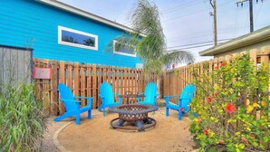 A backyard with a blue house, a wooden fence, a fire pit, and blue chairs.