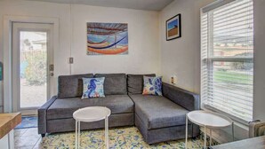 A living room with a grey sofa, two white side tables, a colorful painting, and a window with blinds.