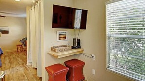 Una habitación con suelo de madera, televisor montado en la pared, ventana con persianas y un pequeño estante con una cesta y un marco de fotos.