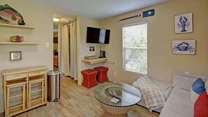 Sala de estar con una mesa de centro de vidrio, un sofá blanco, taburetes rojos, un televisor montado en la pared y una ventana con persianas.