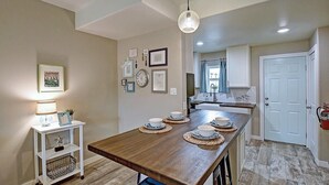 Un comedor con una mesa de madera dispuesta para cuatro, una pared blanca con cuadros enmarcados y una cocina al fondo.