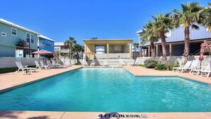 Piscina com espreguiçadeiras e guarda-sóis, cercada por palmeiras e edifícios.