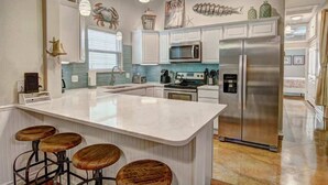 Une cuisine moderne avec des meubles blancs, un grand réfrigérateur en acier inoxydable, un îlot central doté de tabourets de bar et des éléments décoratifs tels qu’une fresque représentant des poissons et des plantes suspendues.