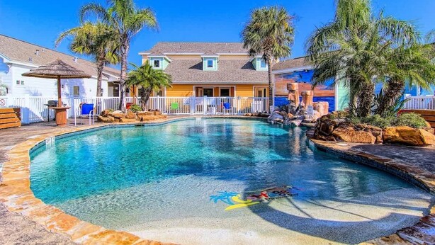 Une piscine à l’eau d’un bleu cristallin, entourée de palmiers et bordée d’un bâtiment à la façade jaune.