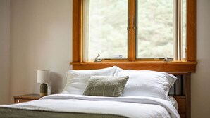 Een slaapkamer met een bed, kussens, een nachtkastje met een lamp en een raam met uitzicht op bomen.