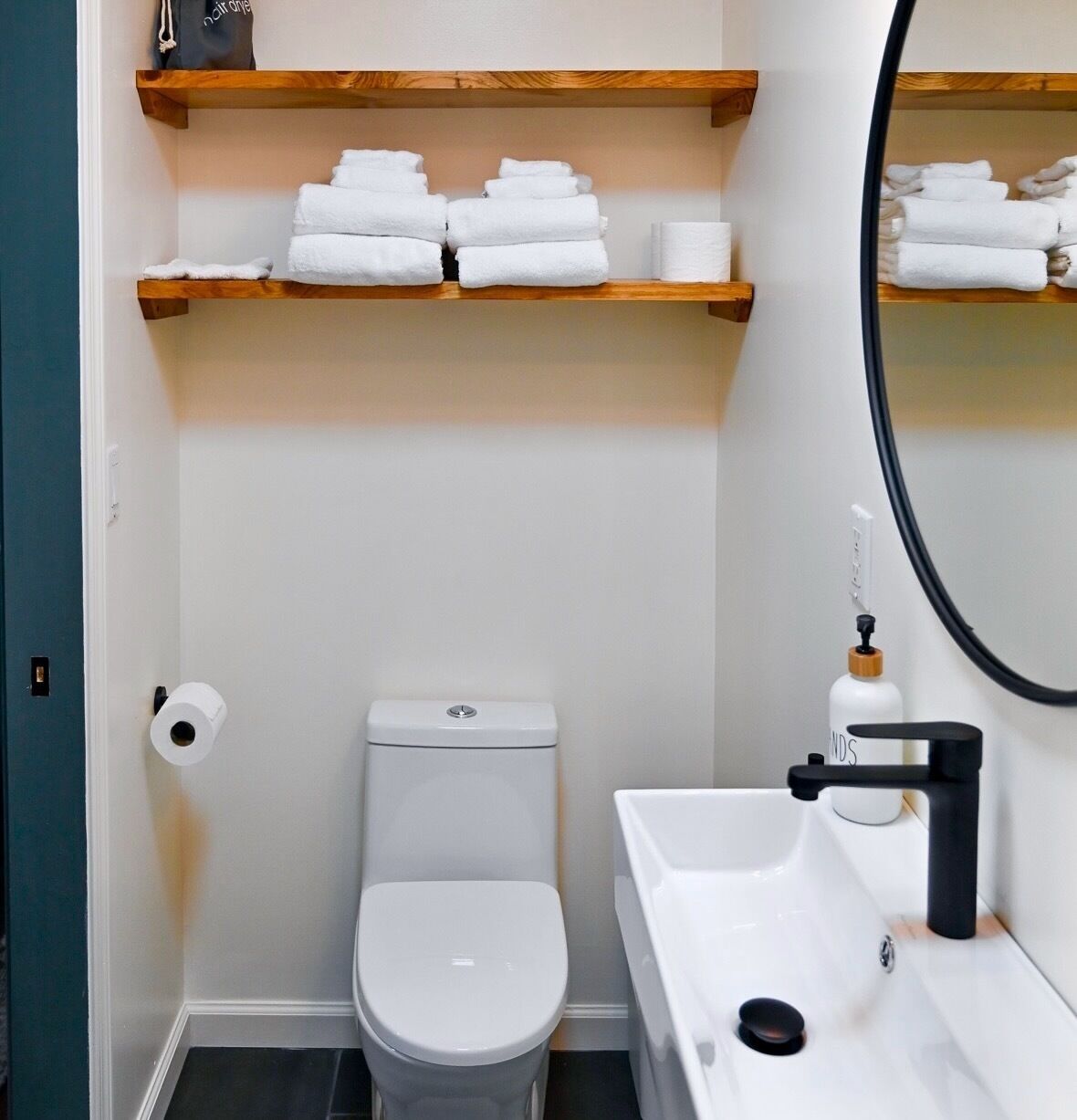 Un bagno compatto con il water, un lavandino e uno specchio. Sopra il water, mensole in legno contengono asciugamani e un rotolo di carta igienica.