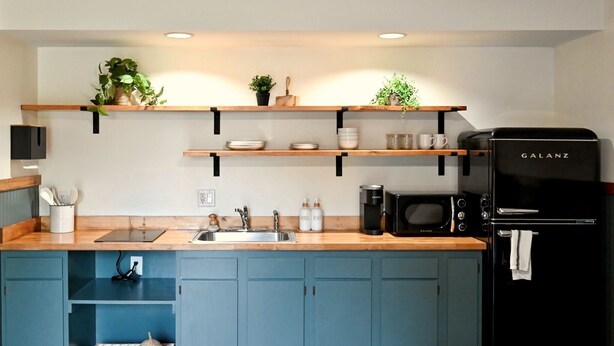 A modern kitchen with a black Galanz refrigerator, a coffee maker, and wooden shelves holding plants and dishes. - The Gate  -  Bungalow (Skaneateles)