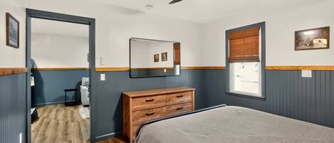 A bedroom with a bed, a dresser, a mirror, a ceiling fan, a window with blinds, and a door leading to another room.