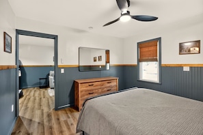 Un dormitorio con cama, cómoda, espejo, ventilador de techo, ventana con persianas y puerta que da a otra habitación.